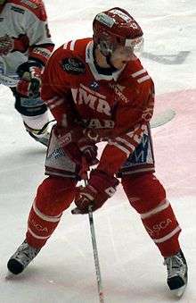 An ice hockey player stands partially crouched, while looking to his left. He is wearing a red helmet and uniform.