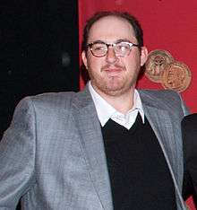 A bespectacled man in a gray suit jacket looks to the camera's left.
