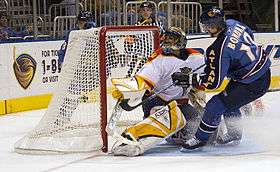 An ice hockey player wearing a blue jersey following through on a shot against a goaltender wearing a white jersey from close proximity. The goaltender's left blocker and pad are outstretched as he watches the puck go in the net behind him.