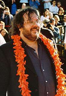 A photograph of Peter Jackson, a brown-haired, bespectacled man wearing a brown blazer, a blue shirt, and an orange lei