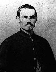 Head and shoulders of a white man with dark hair and a neatly trimmed Van Dyke mustache and beard, wearing a plain dark jacket.