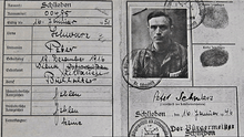 A portion of a small two-page document. On the left is a table with German field names in a Germanic typeface, filled in with handwriting giving names, places and dates. On the right is a black and white photograph of a dark-haired man in a striped top seen from the chest up with a number on the left side at bottom. Next to it are two fingerprints, and below his signature and the date 16 February, 1946. Around this are several stamps.