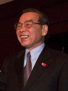 a smiling man with black hair, wearing glasses, a Communist Party insignia, a suit and a red tie