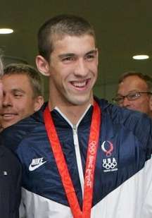 Grinning young man with very short black hair with a gold medal hung around his neck on a red ribbon. He is wearing a white and blue tracksuit designated as the uniform of the United States and someone else's hand is waving an American flag in front of his torso. Two men are standing behind him.