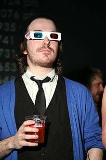 Fish wears his medium-long hair off his face, some stubble facial hair, 3D glasses, a white dress shirt with loosened black tie under a black vest and a blue cardigan while holding a red drink at a party.