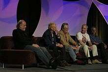 Five older men sit on a couch, with an arched, decorative purple background behind them. Ralston, leaning back in his seat, has short hair and wears a shirt with slacks.
