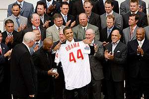 Several rows of men standing and clapping; in the front is a smiling, brown-skinned man holding a white baseball jersey with "Obama" and a large "44" in red on the rear toward camera