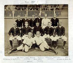 Two rows of men, one standing behind one seated, of men wearing old-style white baseball uniforms and striped pillbox caps