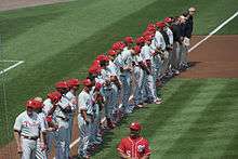 A row of men wearing gray baseball uniforms with red trim and red baseball helmets standing in a line down a dirt path on a grass field