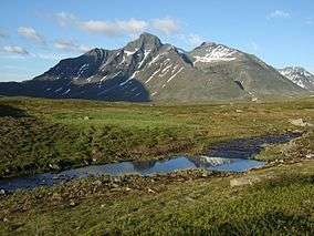Mountain with two main peaks, composed of dark rock with traces of snow on the sides, against a bright blue sky. In the foreground, a stream cuts through grassland.