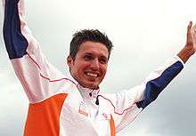 Man with spiky brown hair and stublle raises his arms aloft. He is wearing a white tracksuit top with blue and orange portions.
