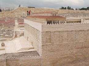 The roof and upper portion of a columned structure rises above a very high stone wall which has wide pilasters decorating the upper part of the wall and a wide square and steps at the foot of the wall.