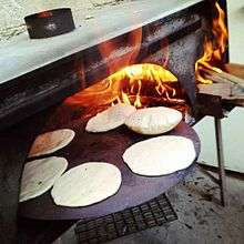 Six pitas baking on a circular pan in a wood-fired oven