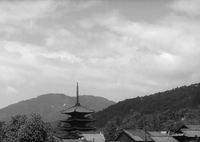 Shot of a Japanese pagoda (in Kyoto), set among ordinary rooftops and trees, with tree-lined mountains and sky in the background.
