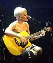 A woman with short blonde hair singing with her microphone while playing a guitar