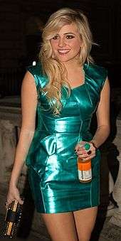 A picture of a woman with blonde hair wearing a green metallic dress holding a bottle of drink.