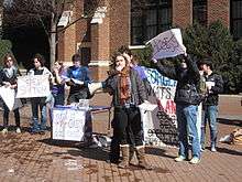 A college-aged female in jacket and scarf holds the microphone attachment of a bullhorn while other students hold protest signs behind her. Two with large red X's over the words read "Free Speech" and "Access."