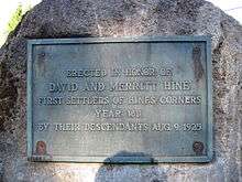 Cast metal plaque bolted to a bolder, showing clear signs of age. The plaque reads, "ERECTED IN HONOR OF/DAVID AND MERRITT HINE/FIRST SETTLERS OF HINES CONRERS/YEAR 1811/BY THEIR DESCENDANTS AUG. 9, 1925."