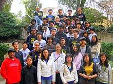 39 students stand on a staircase and face the camera. In the background is foliage.
