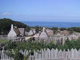 A modern-day photograph of a village consisting of small, primitive wooden houses.  Most of the houses have thatched roofs.  In the distance is a large expanse of ocean and a clear blue sky.  The village is surrounded by a wall consisting of tall, thick wooden planks.