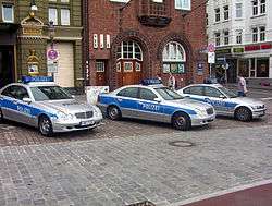 Three patrol cars painted in silver with a blue stripe on the side doors. On the car roof are blue emergency lights.