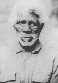 Head and shoulders of a dark-skinned man with bushy white hair and white beard wearing a buttoned-up shirt.