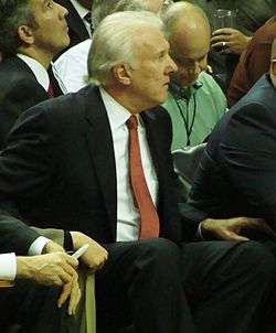 A man with white hair, wearing a black suit, white shirt and orange tie, sitting at a basketball game.