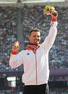 A Caucasian man with dark hair wearing a white zipped up top. He holds a bunch of flowers in his left hand, and clutches a gold medal in his right.