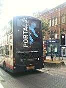 The back of a brown, double-decker bus in a city street, with the text "Portal 2" and a blue symbol of a man that seems to be vanishing through a doorway