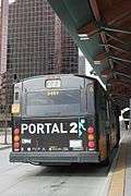 The back of a brown, single-decker bus in a city street, with the text "Portal 2" and a blue symbol of a man that seems to be vanishing through a doorway