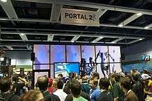 A crowd of people waiting in line in front of a partitioned 20'x20' area within a larger reception hall. The outside of the partition is decorated with Portal 2 artwork.