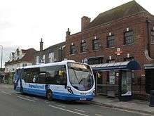 Pale grey bus with blue markings in front of a brick building