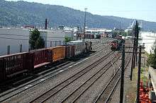 Two trains head in opposite directions along tracks in a switching yard surrounded by warehouses.