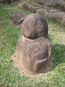 A small stone sculpture with a round featureless head and squat round body. The figure is seen from the front left and the left arm is visible, raised against the side of the figure's body. The arm descends from the shoulder and bends forwards at the elbow, running horizontally to the wrist, which bends sharply downwards, with the fingers marked by carved grooves. The sculpture has no legs and rests on a grassy area in front of some stone steps.