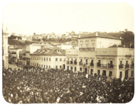 An old photograph showing a crowded square in front of a large, white, multi-storied building