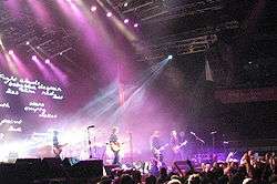 A wide shot of a stage with only four musicians visible: each is playing a type of guitar. A drum kit is mid-stage but the Coghill is obscured by lighting and equipment. The audience are across the front, below stage, some have one fist raised. A photographer is centred, front of stage, another is further to the right with a camera pointing at the band, a third cameraman is at left pointing into the crowd. Behind the group on the left is a large screen which depicts various words including "Fight clouds baby blue despair skin red lies lost stars ith empty rocket paint love g". Overhead lights shine down on the performers. To the right is a large sign with white writing "bring your living room to life" on red-orange background.