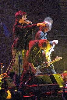 Frontline of Powderfinger are shown in side profile across a stage, with Fanning closest to viewer. He is pointing into the audience with his right fore-finger, while singing into the microphone held in his left hand. Just beyond him, Collins is stooped low over his bass guitar with his long hair fallen forward. Haug is further along and plays his guitar while standing more upright, his shoulder length hair partly obscures his face. Stage equipment is in front of the three men.