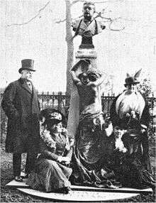 Bond and three other people around an upright outdoor marble base topped by a bust.