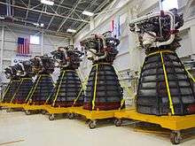 Six rocket engines, consisting of a large bell-shaped nozzle with working parts mounted to the top, stored in a large warehouse with white walls decorated with flags. Each engine has several pieces of red protective equipment attached to it and is mounted on a yellow wheeled pallet-like structure.
