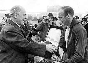 A black and white photograph of two gentlemen. The man to the left, dressed in a dark coat is handing over a trophy inside a box to the man on the right. The man to the right is wearing a dark thin sweater over a lighter sport sweater. Reporters photographing the handover can be seen in the background of the two men.