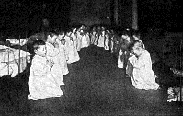 Two rows of little boys, about 20 in total, kneel before their beds in the dormitory of a residential nursery. Their eyes are shut and they are in an attitude of prayer. They wear long white night gowns and behind them are their iron framed beds.
