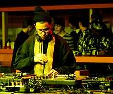 A man wearing eyeglasses, a beanie, scarf, and black sweatshirt, looking down at a set of turntables and other DJ equipment