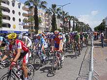 Dozens of professional bicyclists race on a street lined with palm trees and pastel apartments.