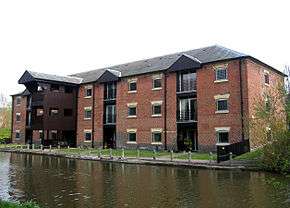 A brick building by the canal with three storeys and a loading bay on the left protruding towards the canal.