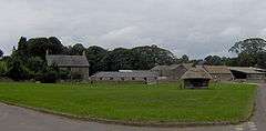 Large area of green grass with stone farm buildings behind. In the centre is a small thatched wooden building.