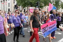 A group of men and women, dressed in purple, pink and blue, marching down a street with a banner which reads "Out and proud - Bisexuals! - Fighting for equality"