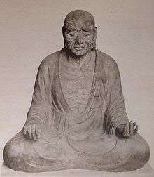 Front view of a cross-legged seated statue with long earlobes. His hands rest on his knees with the palm of his right/left hand turned down/up.