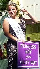 Young woman wearing sleeveless black and white print dress, smiling and waving in a parade. Sign in front of her says Princess Kay of the Milky Way