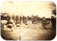 An old photograph showing a procession passing between lines of soldiers with tents in the background