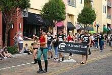 A group of people wearing Pulse logo shirts parade down a brick street carrying a banner with the club's logo on it. An individual dressed as Robin from Batman leads the procession.